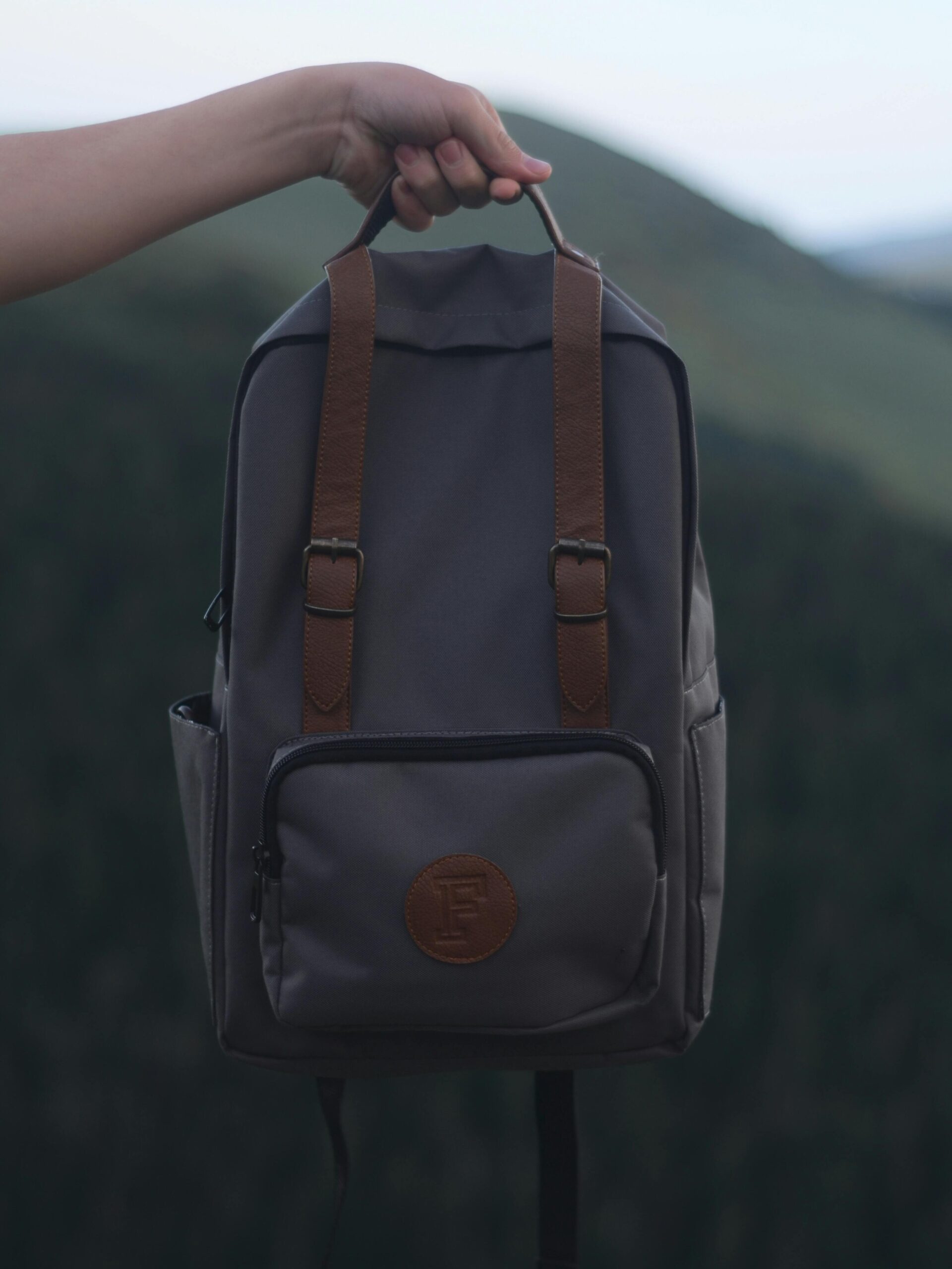 A person holding a stylish gray backpack with leather straps, set against a natural outdoor background.