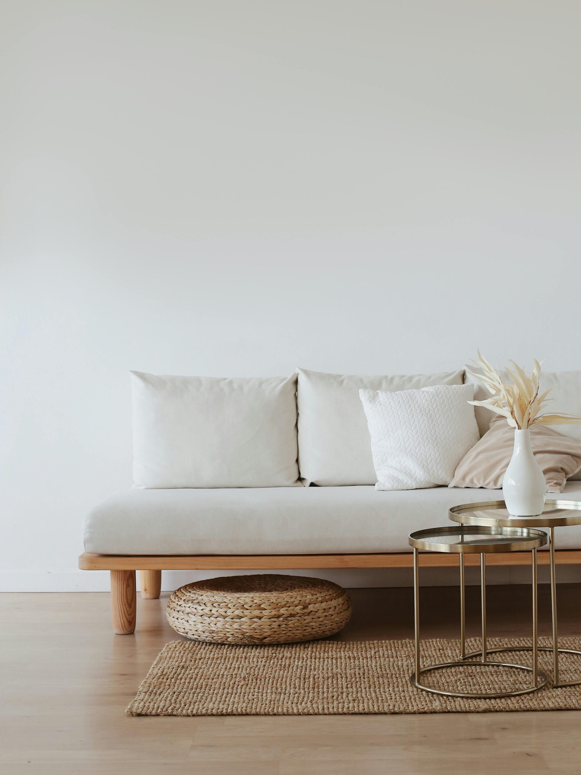 A chic minimalist living room featuring a white sofa with cushions, dried flowers in a vase, and a wooden floor.