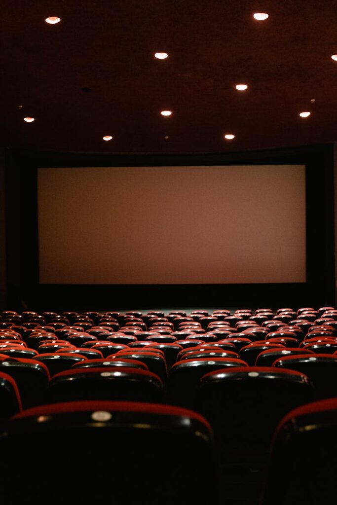 pexels-photo-7991486-7991486 A dimly lit empty cinema theater with rows of red chairs facing a projector screen.